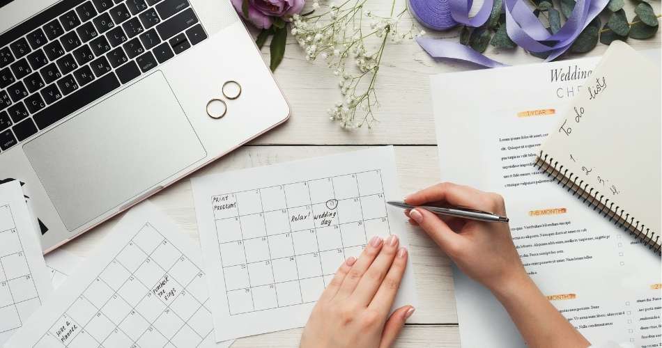 Woman's hands writing notes on Wedding Calendar, two wedding rings are laid on a laptop nearby and a notebook with to do list written on the page is nearby